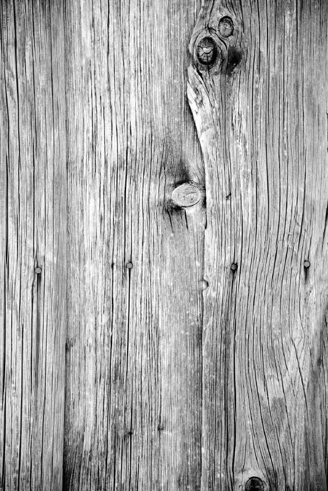 Schwarz-Weiß Bild der Holzfassade an einer Hütte in den Alpen. Man sieht sehr gut die Musterung und an verschiedenen Stellen die Astlöcher und Nägel.