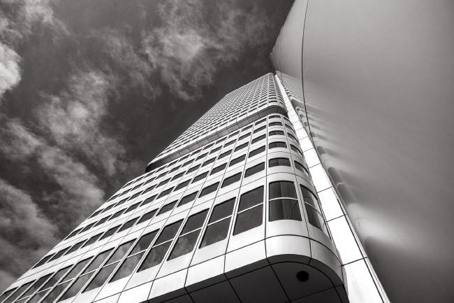 Schwarzweiß Bild im Landscape Format. Der Blick geht leicht schräg auf eine mit Fenstern gespickte Aluminiumfassade die wiederum in Rechteckige Segmente unterteilt ist.. Links oben ist der Himmel mit feinen helleren Wolken zu sehen. Das rechte Drittel des Bildes besteht aus glattem Aluminum das nur einmal unterbrochen wird.