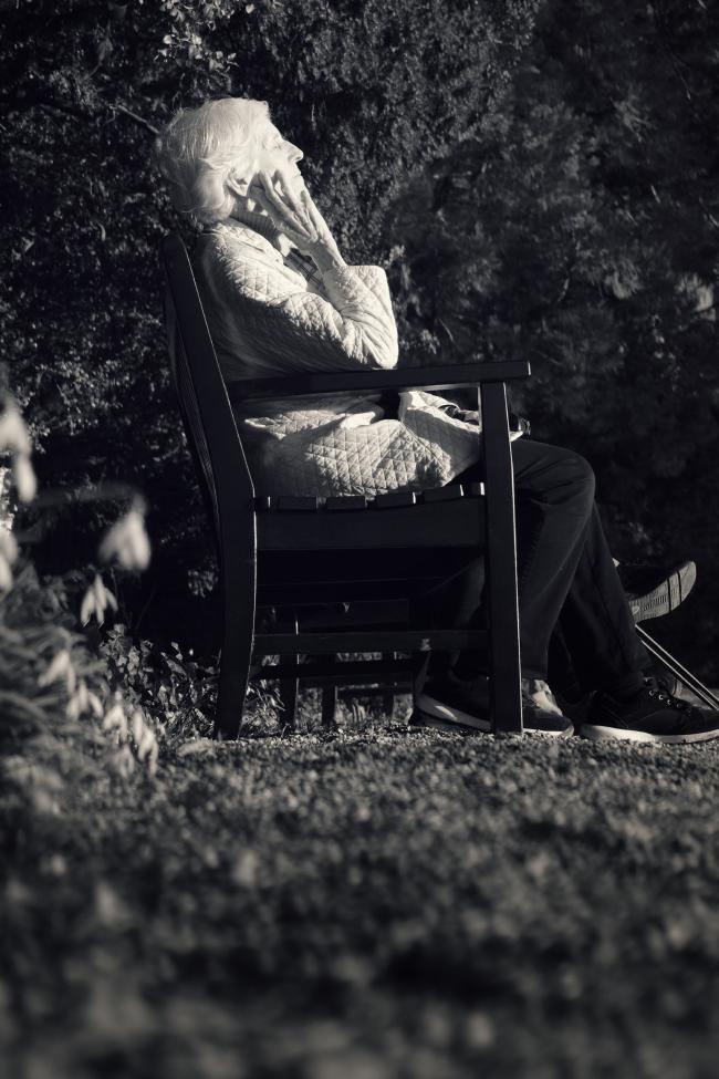 Hochkant Schwarzweiß Bild. Eine ältere Frau mit weißen Haaren sitzt auf einer Bank und genießt die Sonne. Das Bild ist aus einer niedrigen Perspektive aufgenommen und das untere Drittel des Bildes macht unscharfer Kies aus. Links mittig sieht man einige unscharfe Schneeglöckchen. Die Frau trägt eine dunkle Hose und eine helle Jacke mit rautenförmigem Muster. Die Frau hat ihre Hand an ihrer Backe vorm Ohr liegen und genießt mit geschlossenen Augen die Sonne in ihrem Gesicht.