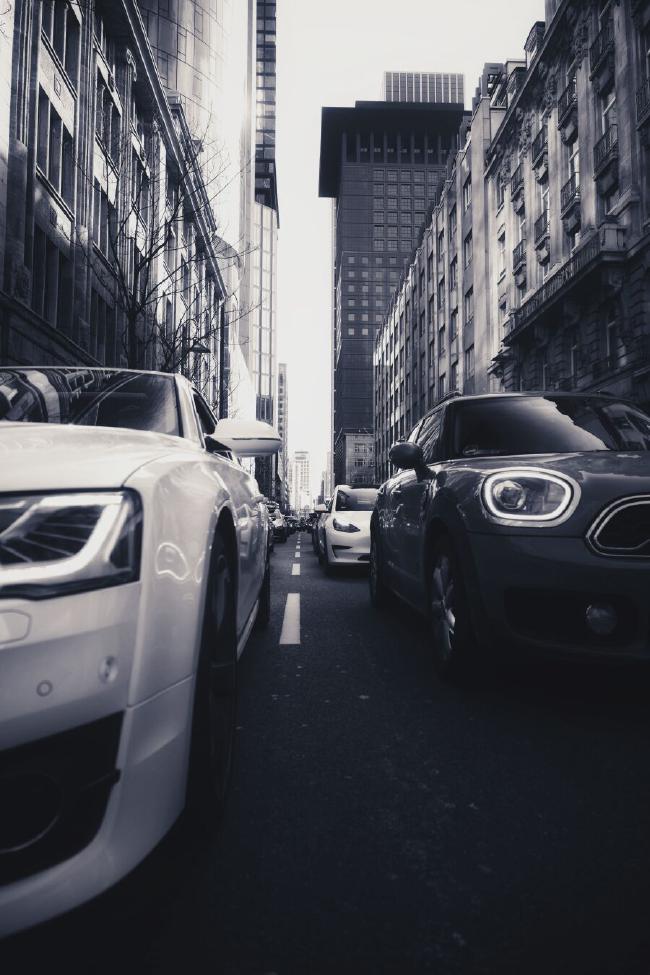 Schwarz-weiß Aufnahme. Man sieht eine Häuserschlucht mit alten Häuserfronten aus Sandstein im Vordergrund und Hochhäusern im Hintergrund. In der Glasfassade des linken Hochhauses spiegelt sich die Sonne. Die Straße ist verstopft mit Autos. Das Bild ist auf Höhe der Frontscheinwerfer in der mitte der Straße aufgenommen, sodass sich eine ungewöhnliche Perspektive ergibt. Der gestrichelte Mittelstreifen ist zu sehen bis er im Hintergrund von noch mehr Autos verdeckt wird die in die enge Straße drängen.