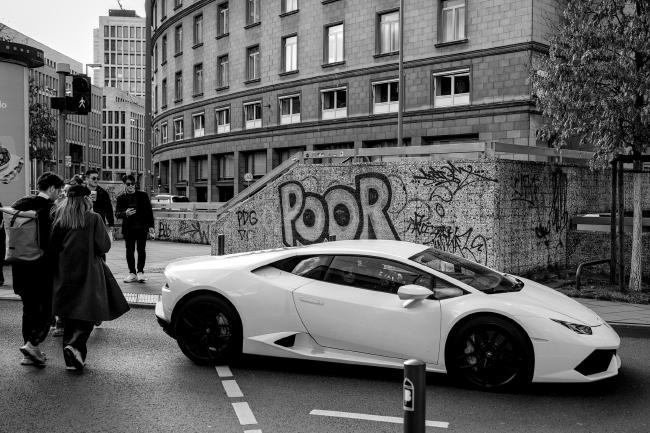 Schwarzweiß Foto im Landscape Format. Zu sehen ist im Vordergrund ein weißer/heller Lamborghini der trotz der grünen Fußgänger Ampel noch teilweise auf dem Überweg steht. Dabei steht er direkt vor einer Wand auf der in großen Buchstaben poor steht. Auf der gegenüberliegenden Seite schauen zwei Männer dem Auto hinterher.