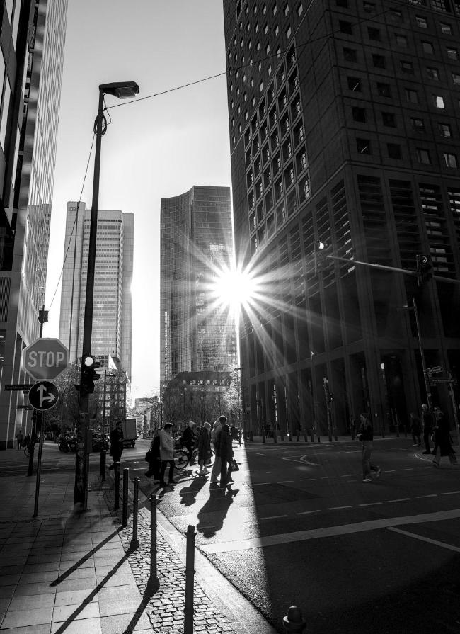 Schwarz-Weiß Bild im Portrait Format. Zu sehen ist ein Straßenübergang auf einer Straße die zwischen einigen Hochhäusern liegt. Diese sind zum Teil nur angeschnitten zu sehen. Das Bild wird von einem Gebäude und seinem Schatten quasi in zwei geteilt und in der Bildmitte ist ein Sonnenstern zu sehen der durch einen optisch feinen Spalt zwischen zwei Hochhäusern scheint. Eine Gruppe an Menschen steht auf der Straße noch im Sonnenlicht und ist kurz davor in den Schatten zu treten.