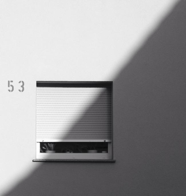 Fast quadratisches Schwarz-Weiß Bild. Zu sehen ist ein Schatten der das Bild in der Mitte diagonal von links unten nach rechts oben teilt. unteren Linkten drittel etwa ist ein Fenster mit fast herunter gelassenem Rollladen und einigen Pflanzen au der Innenseite auf dem Fensterbrett. Daneben steht die Hausnummer 53.