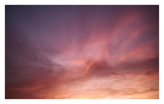 Farbbild im Landscape Format mit weißem Rahmen. Das Bild zeigt sanfte Wolken in den vielfältigen Farben eines Sonnenuntergangs. Während im oberen linken bereich die Blau und Lila Töne dominieren sind es in der Mitte eher Rosa, Rot und Orange. In der rechten unteren Ecke dagegen ein helles Orange und Gelb.