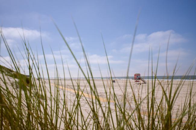 Farbbild im Landscape Format. Blick durch Grashalme an einer Düne auf den Strand und das Meer. Zwischen Düne und Strand verläuft ein Holzweg. Der Himmel ist Blau mit einigen leichten Wolken. Etwas rechts der Mitte findet sich ein strahlend rotes Wachhäuschen der DLRG.