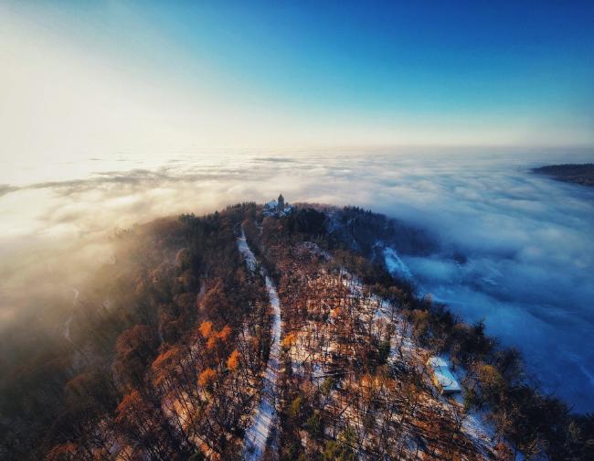Man sieht einen Bergrücken bedeckt mit etwas Schnee zwischen den Bäumen. In der Mitte verläuft ein weg zu einer Burg und das Tal darum liegt im Nebel. Alles ist von der untergehenden Sonne stimmungsvoll angeleuchtet.