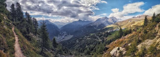 Panorama über das Ötztal bei Sölden von einem Wanderweg aus. In der Mitte und links liegt das Tal eher in Schatten und darüber sind dunklere Wolken. Nach rechts hin wird das Wetter sichtbar besser, sodass der Berg um Hochsölden in der Sonne liegt und am blauen himmel nur einige wenige helle Wolken zu dejen sind. Die Berghänge sind im Tal mit Nadelbäumen bewachsen die bis etwa 60% der Höhe ausdünnen. Im Vordergrund links und rechts sieht man einen Bäume, Büsche und niedrige Vegetation. Rechts verläuft ein dünner Bergwanderweg.