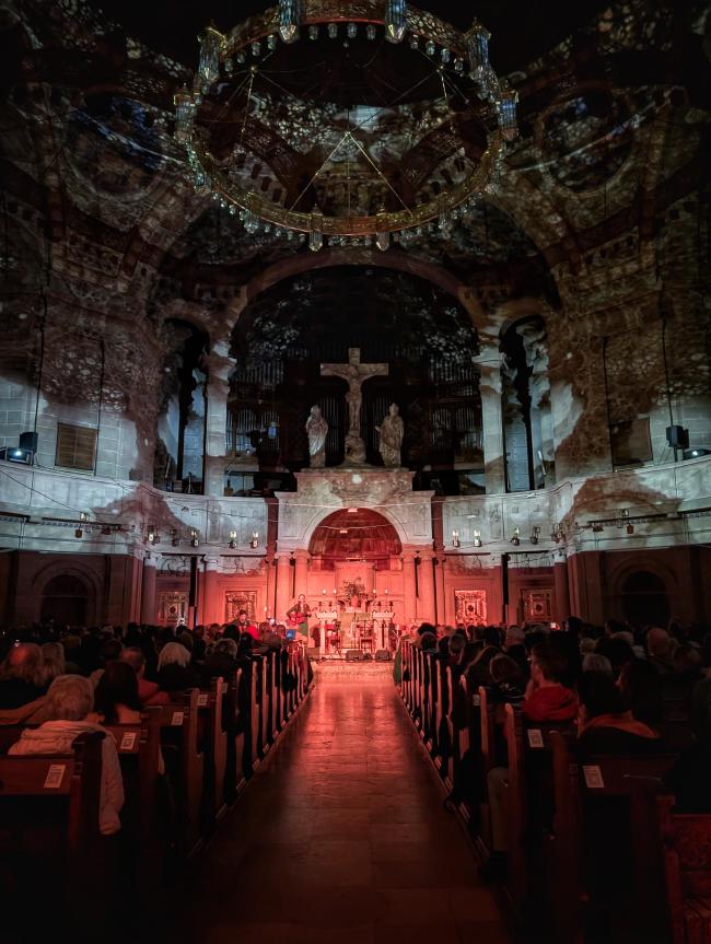 Bild im Portraiformat durch den Mittelgang zur rot beleuchteten Bühne. Darüber sind die Wände und Kuppel in feine Visuals getaucht.