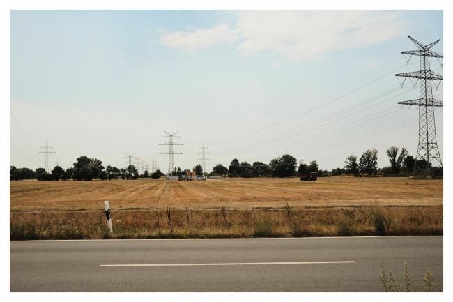 Farbbild im Querformat mit weißem Rahmen. Zu sehen ist im Vordergrund eine leere Straße mit einem Strich in der Mitte. Links auf der gegenüberliegenden Seite steht ein Pfosten. Dahinter ein Gelbes Feld und am Horizont ein paar Dunkle Bäume und Büsche, Strommasten und eine Baustelle. Dahinter sehr hell der diesige blaue Himmel mit ein paar Wolken.