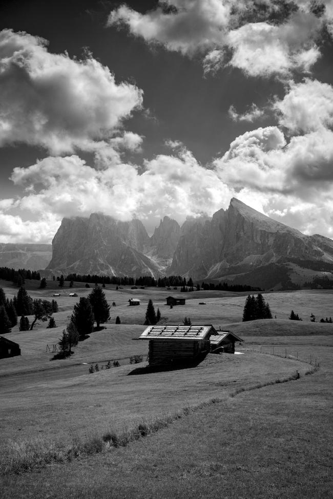 Schwarzweißbild im Portrait Format. Zu sehen ist die sanft geschwungene Landschaft der Seiser Alm in Vordergrund und Bildmitte. Zu erkennen sind dort mehrere Verstreute kleine Hütten, davon eine im Bildvordergrund in der Mitte. Dazwischen befinden sich Wiesen und einige Nadelbäume. Im Bild Hintergrund der etwa in der Bildmitte beginnt ragt das Langkofelmassiv in den Himmel und verschwindet zum Teil in einigen Wolken die den darüber liegenden Himmel bevölkern. In der rechten oberen Bildecke kann man ein paar Sonnestrahlen im Dunst erkennen.