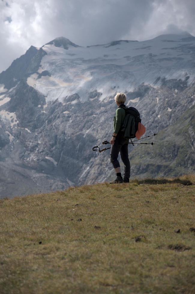 Farbbild, Portrait Format. Man sieht eine Frau mit kurzen blonden Haaren, einer grünen Jacke, grauer Hose, schwarzem Rucksack und Wanderstöcken in der Hand. Sie steht auf einer mit Gras bewachsenen Fläche die hinter ijr abrupt aufzuhören scheint. Weiter hinten im Bild sieht man einen Berg der das ganze Bild einnimmt auf dem oben noch Reste eines Gletschers sind. Darüber der grau bewölkte Himmel.