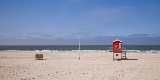 Farbbild im Landscape Format. Ein sehr cleaner Blick von hinten über Strand und Meer. Keine Menschen sind zu sehen. Der Himmel ist Blau mit einigen leichten Wolken am Horizont. Etwas rechts der Mitte findet sich ein strahlend rotes Wachhäuschen der DLRG. In der Mitte ein kreuzförmiger Flaggenmast und links davon ein holzverkleideter Mülleimer. Auf dem Sand sind verschiedene Spuren zu erkennen.