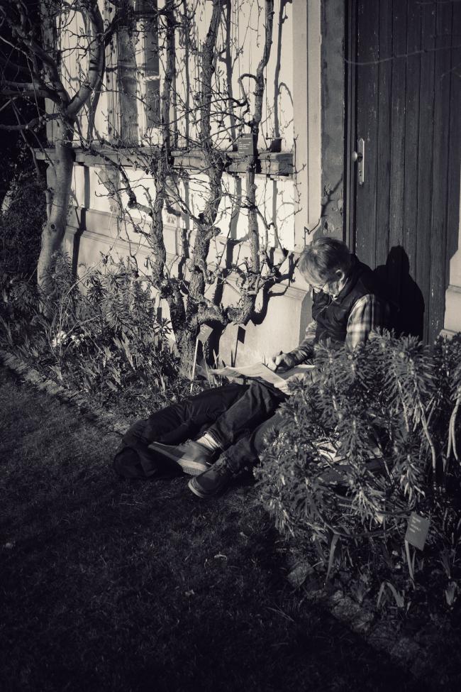 Hochkant Bild in Schwarzweiß. Eine Person sitzt vor einer Holztüre eines älteren Gebäudes, schaut in eine Zeitung und macht dort irgendetwas mit einem Stift. Vielleicht ein Kreuzworträtsel. Die Person trägt eine dunkle Hose, ein Kariertes Hemd und eine Schwarze Weste. Die Person hat kurze Haare und trägt eine Brille. Die Person ist vermutlich zwischen 50 und 70 Jahre alt. Links und recht neben der Person sind Beete mit Pflanzen. Im Linken Beet wachsen Pflanzen die sich an Halteseilen nach oben Ranken und Blattlos Schatten auf der hellen Fassade hinterlassen. Die Linke untere Ecke macht Wiese aus die schon im Schatten liegt und die sich vor der Person und den Beeten befindet.