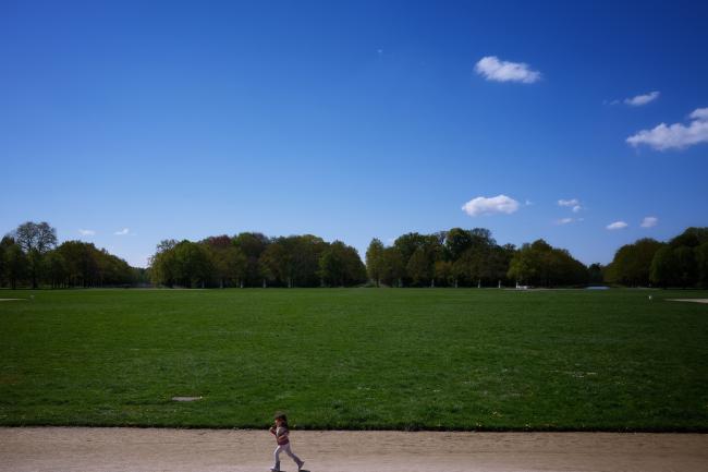 Ein kleines Kind rennt auf einem Weg vor einer riesigen grünen Wiese an deren Ende Bäume stehen und sich links und rechts symmetrische Wasserbecken erstrecken. Es sind wenn man nicht sehr sehr genau guckt keine anderen Menschen zu sehen und der Himmel ist fast wolkenlos. 