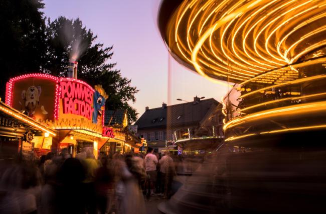 Farbbild im Landscape Format. Das Bild ist eine Langzeitbelichtung wodurch die sich durch die Gegend schiebenden Menschenmassen unscharf sind. Links sieht man einen großen Verkaufsstand auf dem in großen roten Lettern 'Pommes Factory' steht. Rechts sieht man die Kreise von Lampen von einem großen Etagenkarussell. Die Lichtstimmung ist ein leicht rosa blauer Himmel vom Sonnenuntergang.