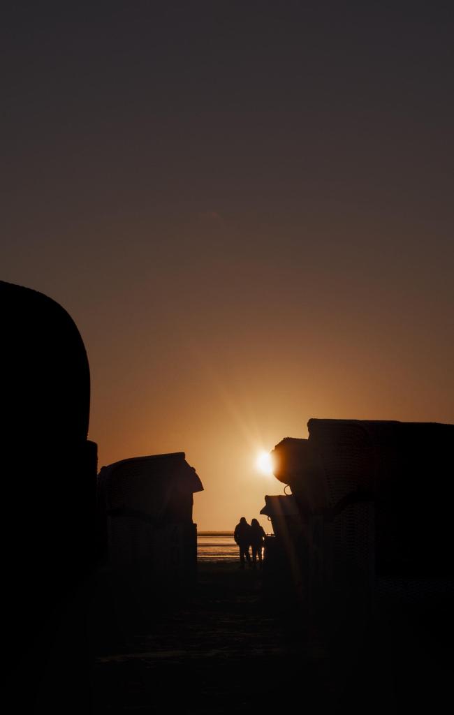 Farbbild im Portrait Format. Zu sehen sind die schwarzen Shiluetten von Strandkörben die eine Art Durchgang bilden an dessen Ausgang zwei Personen Arm in Arm stehen. Hinter diesen ist klein ein dünner Streifen Meer zu sehen. Die oberen zwei Drittel des Bildes sind der wolkenlose Himmel der in Orangetöne getaucht ist und hinter einem der Strandkörbe scheint mittig leicht die Sonne hervor.