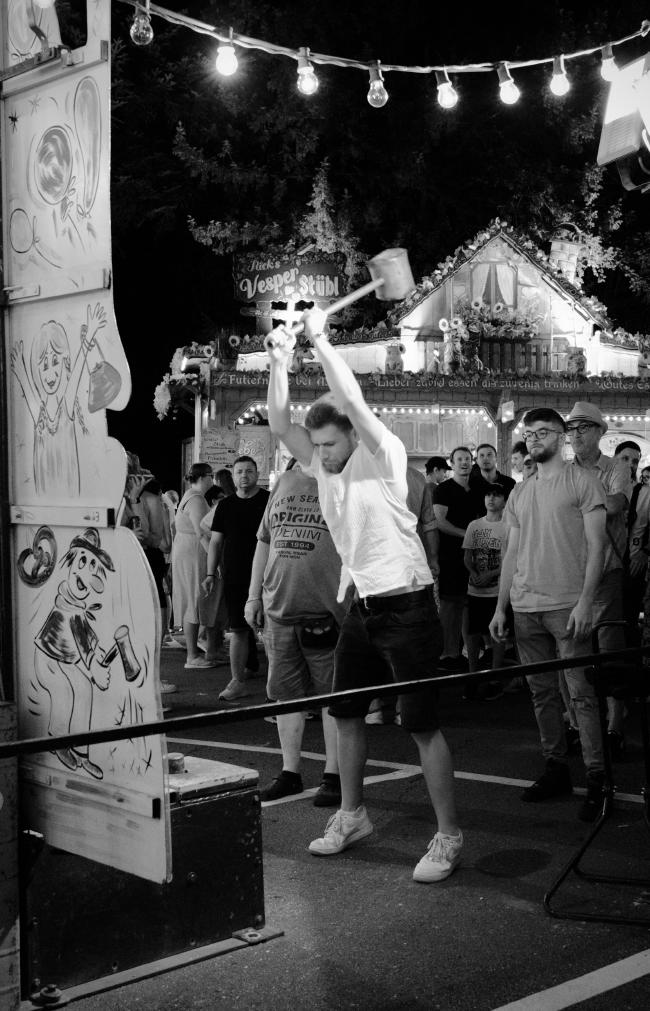 Schwarzweißbild im Portrait Format. Ein Mann in einem weißen Hemd schwingt gerade einen großen Hammer über seinem Kopf. Das Gesicht ist Angestrengt und im Hintergrund stehen einige Zuschauer. Links im Bild sieht man zum Teil den Hau den Lukas auf den er gleich schlagen wird. Im oberen Bildbereich sieht man eine Lichterkette und im Hintergrund Eine beleuchtete Hütte mit Bäumen dahinter.