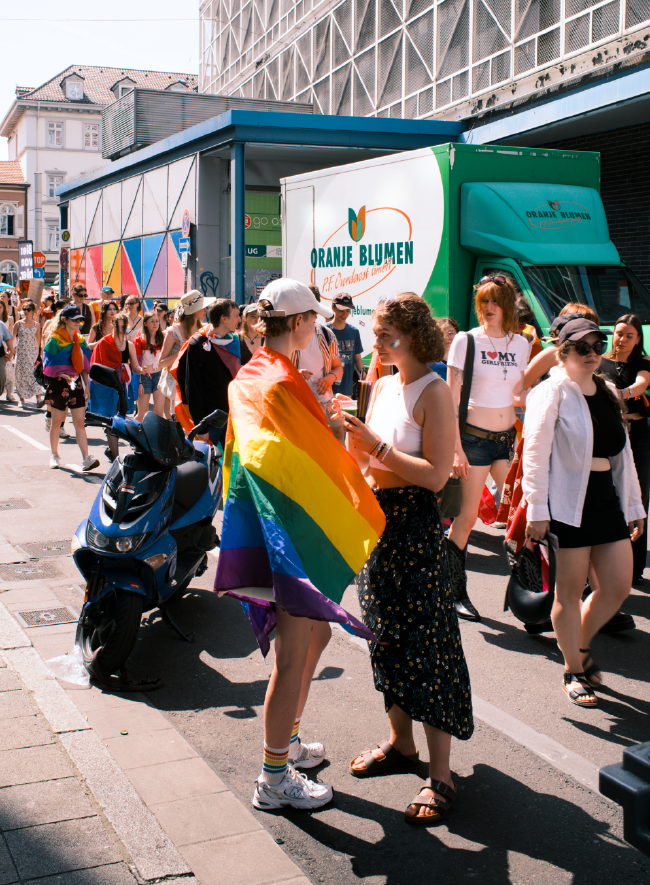 Farbbild am Rande des CSD. Zwei Personen stehen sich gegenüber und haben einen direkten Blickkontakt. Eine davon hat eine Regenbogenfahne über die Schulter. Hinter ihnen laufen die anderen CDS Teilnehmenden an ihnen vorbei. Links neben den zwei Personen steht ein Mopped und hinter der Demogruppe ist ein LKW von Oranje Blumen zu sehen der gerade an einer Laderampe steht.