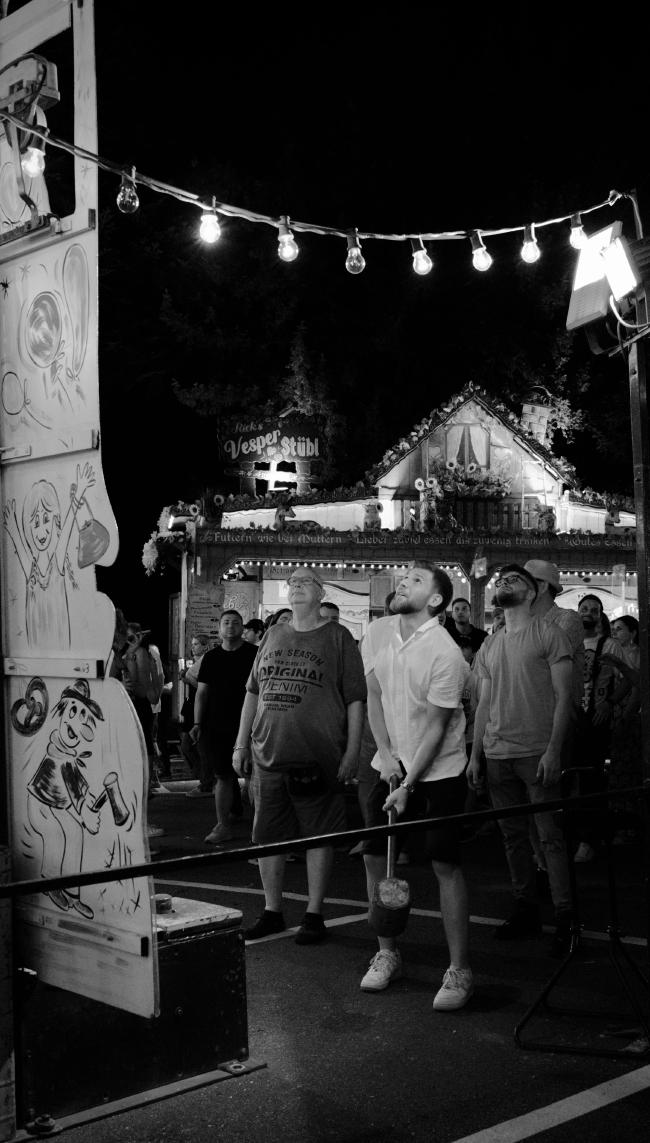 Schwarzweißbild in einem 2/1 Portrait Format. Ein Mann in einem weißen Hemd hält einen großen Hammer in der Hand und schaut Hoffnungsvoll nach oben auf sein Ergebnis beim Hau den Lukas. Im Hintergrund stehen einige Zuschauer und schauen ebenfalls nach oben. Der Betreiber, links vom Spieler schaut dabei unbeeindruckt. Links im Bild sieht man zum Teil den Hau den Lukas. Im oberen Bildbereich sieht man eine Lichterkette und im Hintergrund Eine beleuchtete Hütte mit Bäumen dahinter.