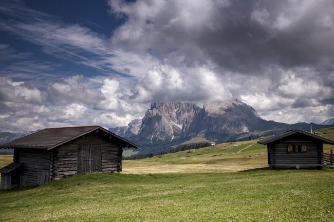 Farbbild im Landscape Format. Der Blick führt zwischen zwei kleine Almen Hütten hindurch über grüne leicht geschwungene Wiesen hin zum Langkofelmassiv, das Mittig in der Ferne liegt. Der Himmel ist teilweise von größen Haufenwolken bedeckt.