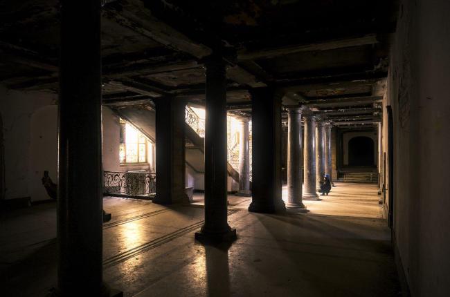 Farbbild im Landscape Format. Das Bild ist aus der Ecke des Raums aufgenommen dessen Maße nicht klar erkennbar sind. Der Raum wird aber durch viele Säulen gestützt. Etwas entfernt in der Bildmitte hinter einigen Säulen sieht man teilweise zwei Treppen die Links und rechts ins obere Stockwerk führen. Dahinter scheint Sonnenlich durch die großen Fenster und flutet den ansonsten dunklen Raum teilweise mit Licht. Eine Person kniet auf der Rechten Seite vor der Mitte des Treppenaufgangs auf dem Boden und macht ein Foto.
