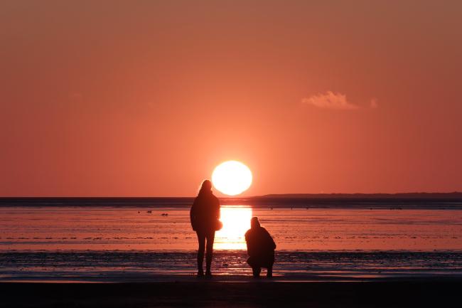 Farbbild in Landscape Orientierung. Zwei Personen am Strand die in den Sonnenuntergang sehen. Eine der Personen steht und eine ist in der Hocke. Die beiden stehen jeweils links und rechts von einem Band Sonnenlicht das sich auf dem Nassen Watt abzeichnet. Die Sonne ist dabei nur noch minimal davor mit der Unterkante den Horizont zu treffen. Auf dem Watt kann man links ein paar Vögel erkennen. Der Himmel is Wolkenlos bis auf eine kleine fast transparente Wolke rechts im Bild. Am Horizont ist der flache Ausläufer einer Insel zu erkennen. Der Himmel und auch das reflektierende Nass ist in ein tiefes orange rot getaucht.