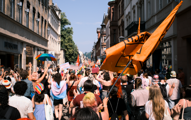 Farbbild. Bild über die Demonstration hinweg durch die Heidelberger Innenstadt. Man sieht viele Menschen mit verschiedenen Flaggen oder anderen Dingen in verschiedenen Pride Farben. Rechts im Bild ist eine große orange aber zusammengefallene Kein Mensch ist Illegal Flagge zu sehen.