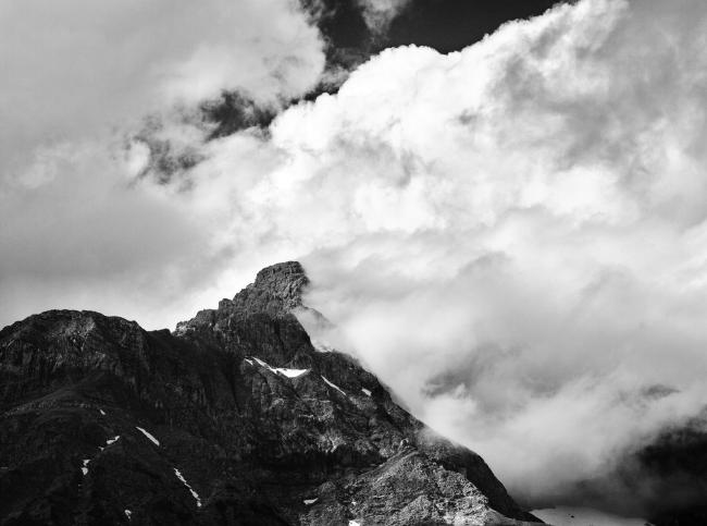 Berggipfel umgeben von Wolken. Auf einer Seite ziehen sich die Wolken vom Gipfel weg. Das ganze Bild ist Schwarz-Weiß.