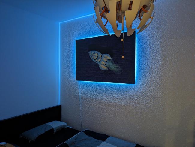 Bild von einem Bild an der Wand über einem Bett das Blau von hinten beleuchtet ist.