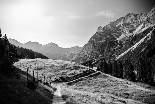 Schwarz-Weiß Bild. in der Mitte des Bildes befindet sich ein Weg der leicht nach rechts zur Bildmitte hin abknickt und etwas links von der Bildmitte startet. Rechts vom Weg sieht man schroffere Berge die im unteren Teil noch von Bäumen bedeckt sind. Rechts sieht man einen abgezäunte Weide auf einem Hügel und am oberen Rand dessen Büme und ein Haus in den Schatten. Im Hintergrund sieht man weitere Berge. Der Himmel ist fast wolkenlos. Das Licht kommt im Bild von schräg Links oben, aber noch recht flach was den Eindruck von morgendlichem Licht im Bild erzeugt.