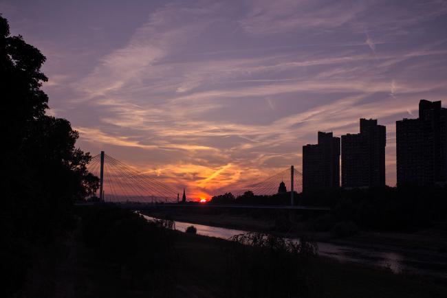 Bild von einem Sonnenuntergang über den Neckar. In der Bildmitte sieht man die Tragseile einer Fußgängerbrücke und rechts drei Hochhäuser.