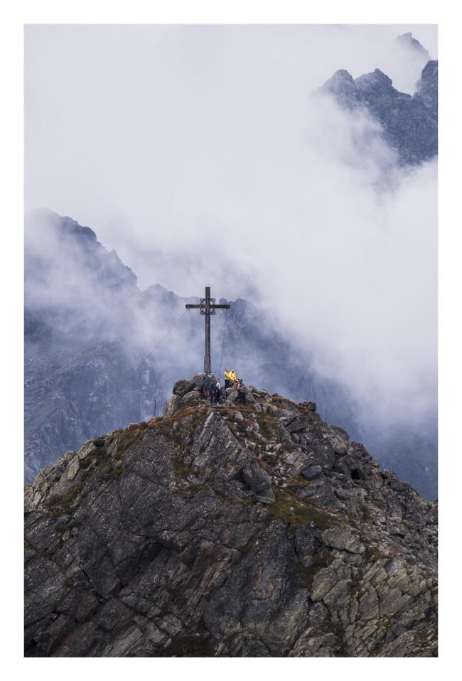 Farbbild im Hochkant Format mit weißem Rahmen. Zu sehen sind ein paar Menschen auf einem Gipfel mit Gipfelkreuz, dahinter sehr komprimiert Wolken und weitere Gipfel die daraus hervorschauen. 