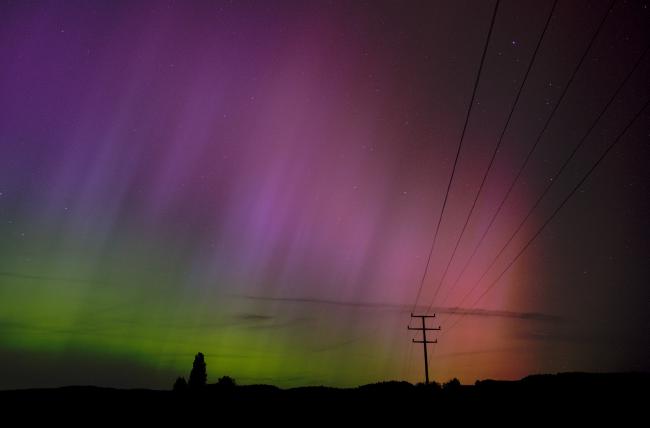 Es sind wellenartige rötlich über lila bis grüne Polarlichter zu sehen. Im Hintergrund liegt der Odenwald und den Vordergrund durchzieht ein Strommast mit seinen Leitungen