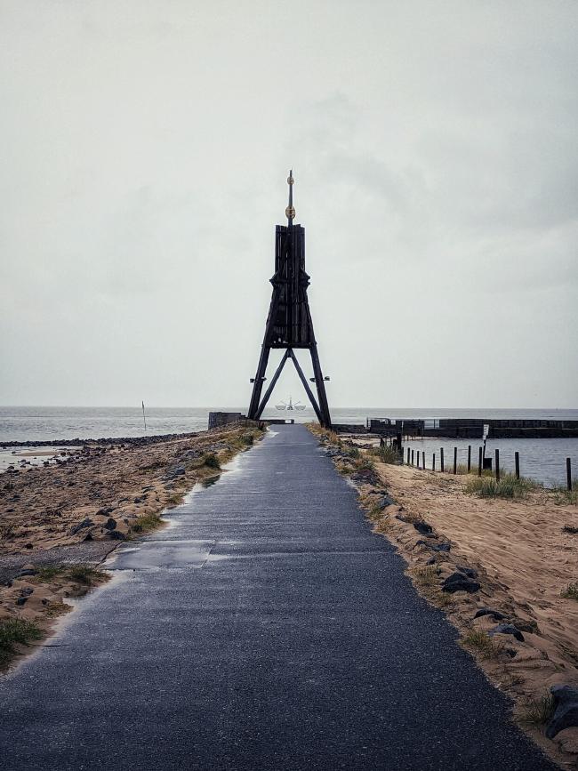 Ein weg führt auf eine große Holzkonstruktion zu. Links und rechts ist Sand und Steine. Im Hintergrund ist Meer. Zwischen den Beinen der Holzkonstruktion sieht man einen Fischkutter mit ausgeklappten Netzen 