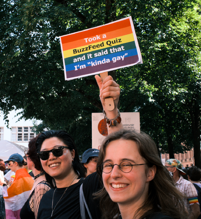 Zwei Personen lachen in die Kamera. Im Hintergrund sind ein paar Bäume, eine Fassade und noch ein paar Menschen zu erkennen. Die Hintere der zwei Personen hält ein Schild in Regenbogen Farben hoch auf dem steht: Took a BuzzFeed Quiz and it said that I'm kinda gay.