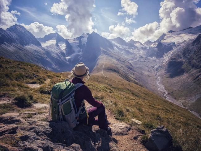 Farbaufnahme im Landscape Format. Eine Person mit roter Jacke, lila Hose, einem Hut und einem großen grünen Rucksack sitzt von der Kamera abgewandt auf einem Stein und guckt in die Ferne. Der Blick führt über einen mit Gras bewachsenen Bergsattel mit Tälern links und rechts. Im rechten Tal sieht man einen Flusslauf. Im Bild Hintergrund erheben sich hohe Berge mit Teilen von Gletschern. Darüber hängen einige Quellwolken am sonst blauen Himmel. Man kann richtig die Weite und Größe der Landschaft spüren wenn man das Bild betrachtet.