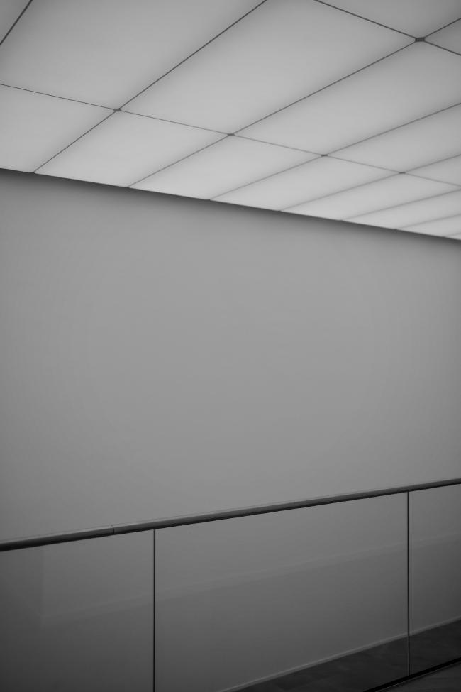 Schwarzweißbild im Portrait Format. Durch das untere Drittel verläuft eine Brüstung aus Glas mit einem oberen Abschluss aus Edelstahl. In der Mitte und durch die Glasbrüstung sieht man eine einfache leere graue  Wand. Darüber befindet sich im oberen Drittel eine aus großen rechteckigen Flächen im 3:1 Format fast ohne Rand bestehende Beleuchtung. in der unteren Rechten Ecke kann man durch die Brüstung noch den Boden des Raums unter der Brüstung erahnen. Alles wirkt sehr clean.