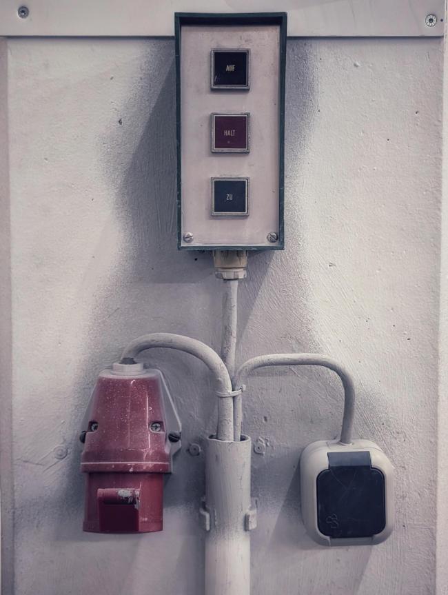 Relativ symmetrisches Bild in Farbe und Portrait Format. Man sieht drei Kabel die am unteren Bildende aus einem weißen Rohr kommen. Zwei Kabel der selben Farbe gehen nach links und rechts und knicken mehr oder weniger im 90° Winkel ab. Einmal in eine Schuko Feuchtraum Steckdose und einmal in eine rote 3 Phase CEE Dose. In der Mitte geht ein weiteres Kabel zu einem Schalter mit den Knöpfen Auf, Halt und Zu untereinander mittig auf einer Edelstahlplatte. Das alles ist auf einer weißen Wand Montiert.