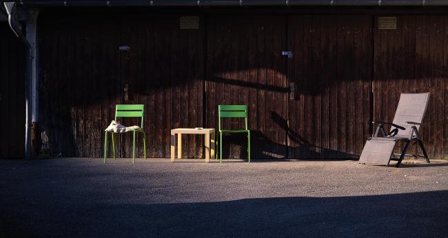 Farbbild ca. im 2:1 Panorma Format. Vor einer alten Garage mit Holztoren stehen etwa Bildmittig zwei grüne Stühle und ein kleiner weißer Ikea Lack Tisch dazwischen. Am rechten Bildrand steht ein grauer Liegestuhl. Die ganze Szene ist von Links von der untergehenden Sonne beleuchtet. Unten und oben ist das Bild im Schatten.