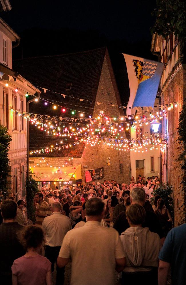 Farbbild im Portrait Format. Eine große menge Menschen, alle in der Nähe der Kamera schauen von der Kamera weg, stehen in einer Gasse zwischen alten Fachwerkhäusern zwischen denen Wimpel und bunte Lichterketten gespannt sind. Rechts oben weht eine Fahne mit dem Weinheimer Stadtwappen.