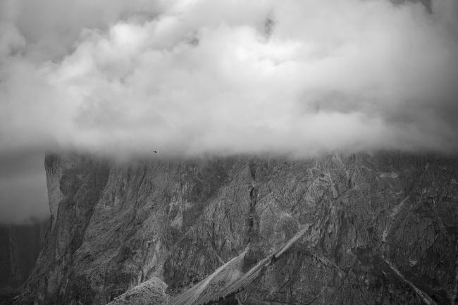 Schwarz-Weiß Bild im Landscape Format. Die untere Hälfte wird von einer Bergflanke dominiert, deren Ende man an der Linken Bildseite sehen kann. Der Berg verschwindet jedoch ab der Hälfte des Bildes in Wolken und gerade an dieser Grenze leicht rechts der Mitte fliegt ein Hubschrauber.