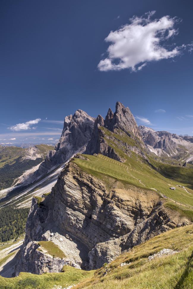 Farbbild im Portrait Format. Man sieht die abrupt abbrechenden Hänge der Seceda in den Dolomiten die oben bis zum Rand mit Gras bewachsen ist, das Muster vom Mähnen zeigt. Dahinter erheben sich die Gipfel der Geisler Gruppe. Rechts im Bild sind klein ein Wanderweg und ein paar Hütten zu sehen,