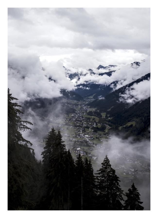 Farbbild im Hochkantformat mit weißem Rahmen. Hier sieht man den Blick aus der Gondel. Im Vordergrund ein paar dunkle Nadelbäume, dahinter zwischen Wolken das Tal mit Straße und Häusern und am Rand jeweils die Berge die zum Teil gerade so aus den Wolken gucken.