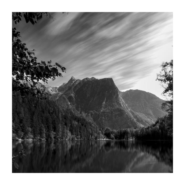 Quadratisches Schwarz-Weiß Bild von einem See gerahmt von einzelnen Ästen und Blättern von Bäumen. Im Hintergrund sticht ein Berg mit seiner Markanten Form in der Bildmitte heraus. Darüber sieht man durch die Langzeitbelichtung die bewegungsunscharfen dahinziehenden Wolken.