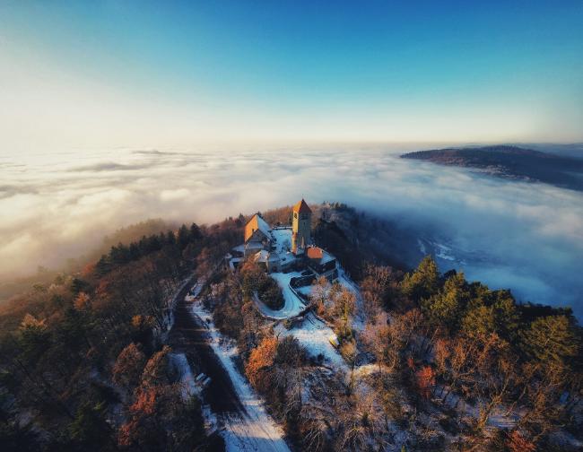 Bild einer schneebedeckten Burg. Die Täler darum sind mit Nebel bedeckt. Alles im Licht der untergehenden Sonne.