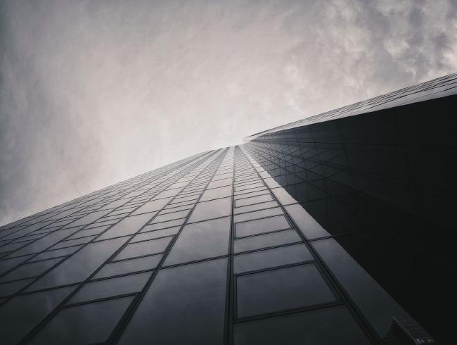 Schwarz-weiß Aufnahme einer Hochhaus Glasfassade. Das Gebäude nimmt etwa die untere Hälfte des Bildes ein und bildet eine Linie von links unten nach rechts oben. Alle anderen Linien bewegen sich zu einem Fluchtpunkt in der Mitte. Der Himmel ist gräulich mit etwas Struktur von einer dünnen Wolkendecke.