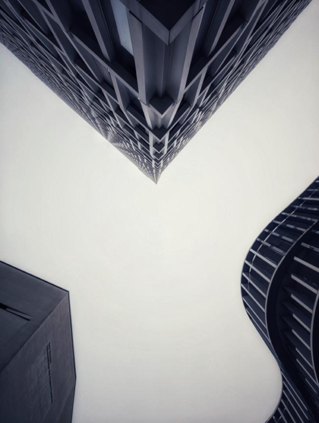 Relativ monochromes Bild im Portrait Format. Darauf zu sehen sind drei Gebäude von unten Richtung Himmel fotografiert. Das erste das von oben in Form eines Dreiecks Richtung Mitte ins Bild schaut hat relativ viele Betonstrukturen die aber relativ geordnet wirken. Rechts unten ist ein Gebäude mit geschwungener Wellen-Form und Parallelen Trägern orthogonal zun Himmel. Und links unten ein Gebäude mit einer sehr simplen glatten Fassade und einer eher angeschnitten trapezförmigen Form.
