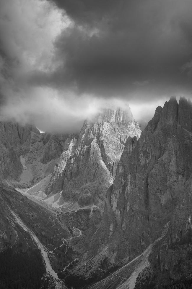 Schwarzweißbild im Portrait Format. Zu sehen ist das innere des Langkofelmassivs. Der Pollice? steht dort mittig und wird zwischen den ansonsten dichten und dunklen Wolken von der Sonne angestrahlt. Zwoschen den Felsen der Berge sieht man einen Weg sich nach oben schlängeln.