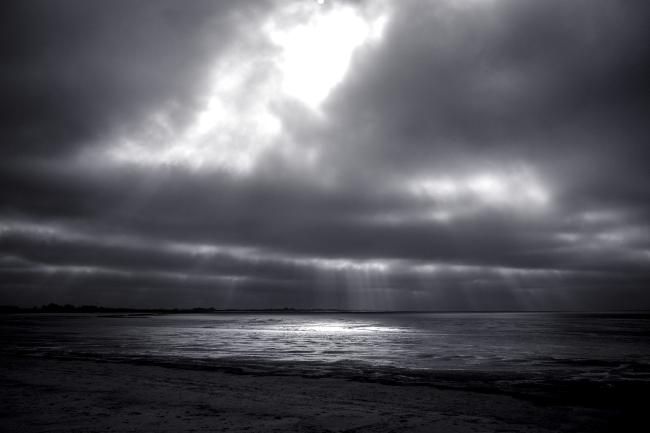 Schwarz-Weiß Bild. Blick über das Watt bei Ebbe. Es ist stark bewölkt aber es scheinen einzelne sichtbare Lichtstreifen durch die Wolken auf das Watt wo sie sich spiegeln. Hier ist es eher ein großes Licht.