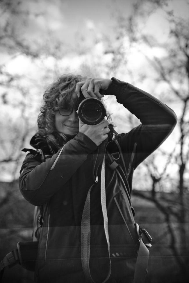 Bild von Leah in einer spiegelnden Scheibe die Kamera vorm einen Auge, sodass nur das halbe Gesicht zu erkennen ist.